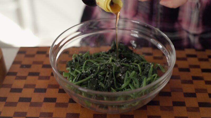 Sesame oil is added to seasoned spinach in a bowl.