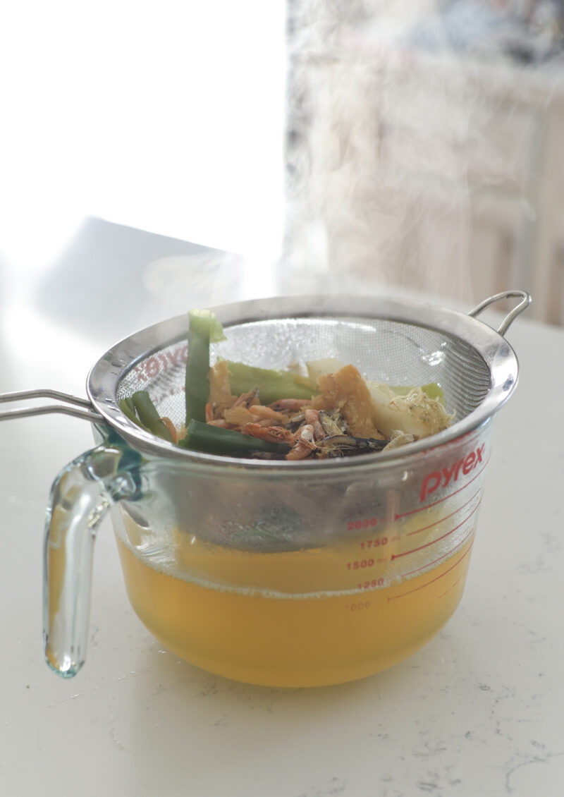 A mesh strainer over a glass bowl is holding the debris of stock and reserving the liquid.