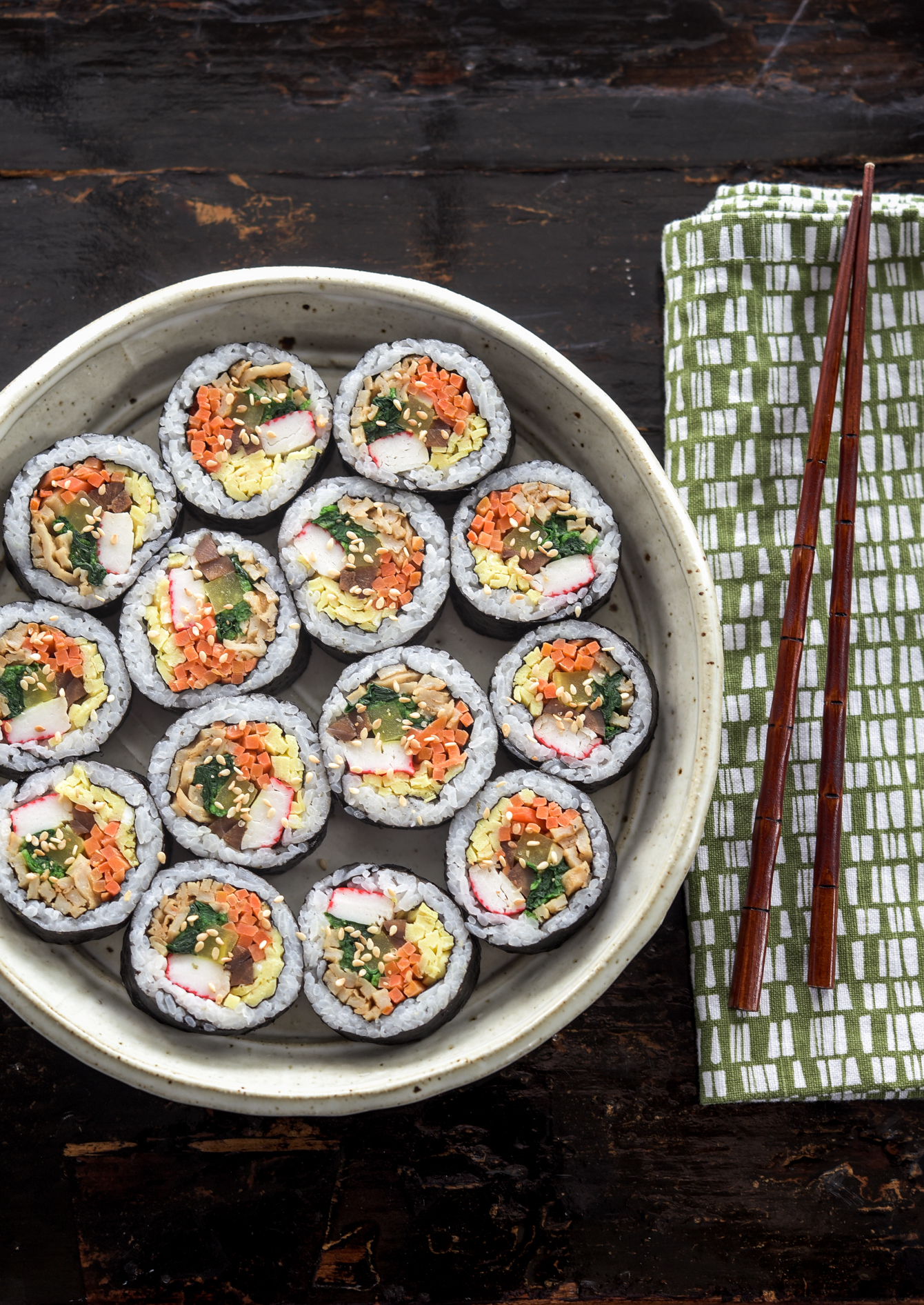 Slices of colorful kimbap with vegetable filling is served on a round plate with a pair of chopstick