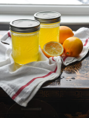 Two jars of Meyer lemon syrup are displayed with lemons on the white kitchen clothe.