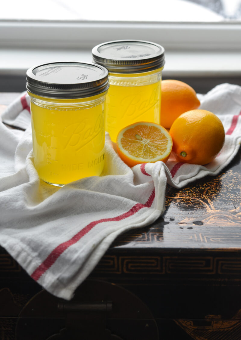 Two jars of Meyer lemon syrup are displayed with lemons on the white kitchen clothe.