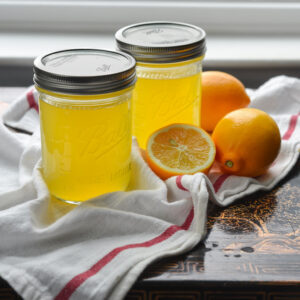 Two jars of Meyer lemon syrup are displayed with lemons on the white kitchen clothe.