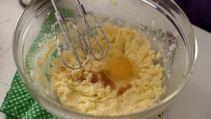 An egg and vanilla extract is added to creamed butter in a bowl.