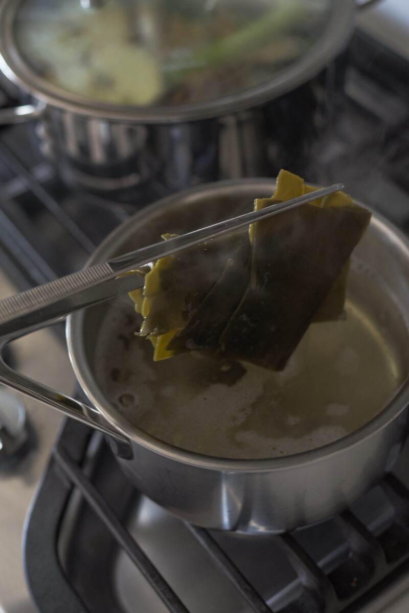 Kitchen tongs are removing the sea kelp from the simmering stock in a pan.