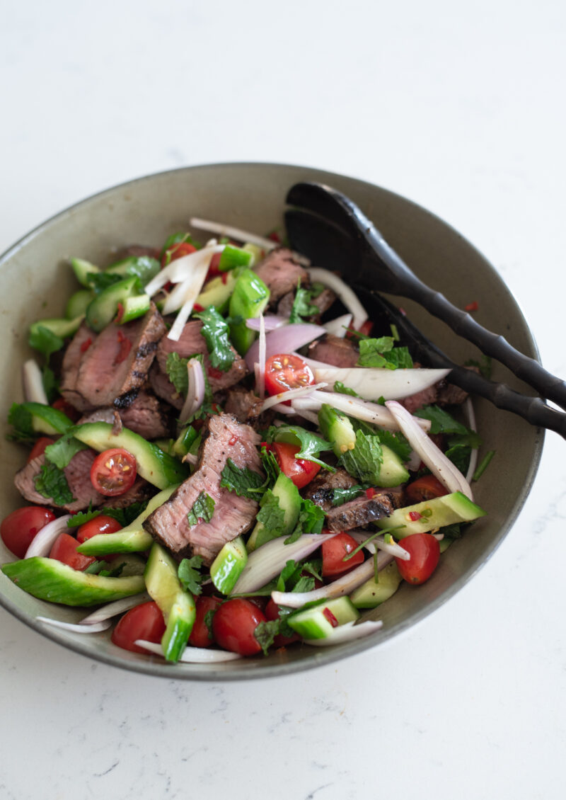 Seared beef streak, cucumber, tomato, and herbs are combined in a bowl for Thai beef salad