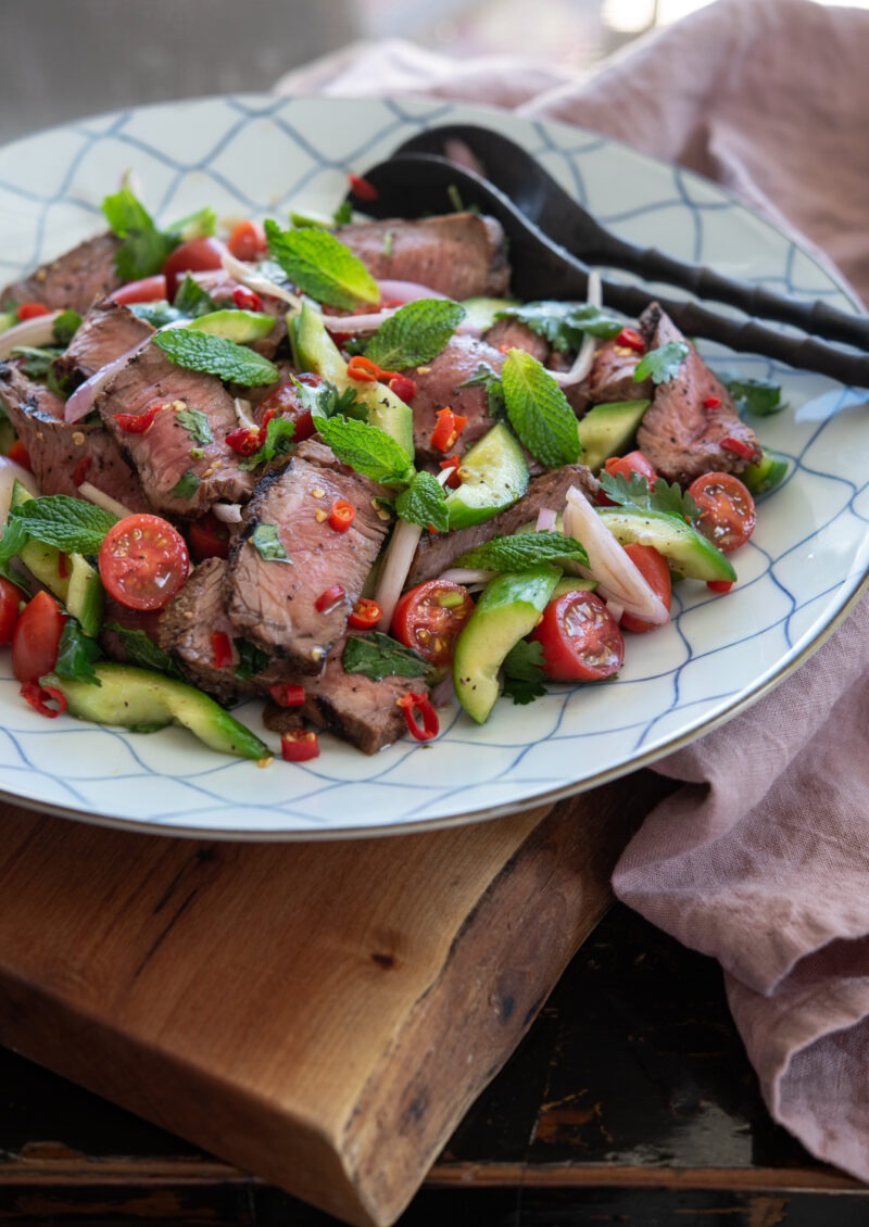 A large serving plate of Thai beef salad (Yum Nua) with cucumber and tomato is garnished with herbs.