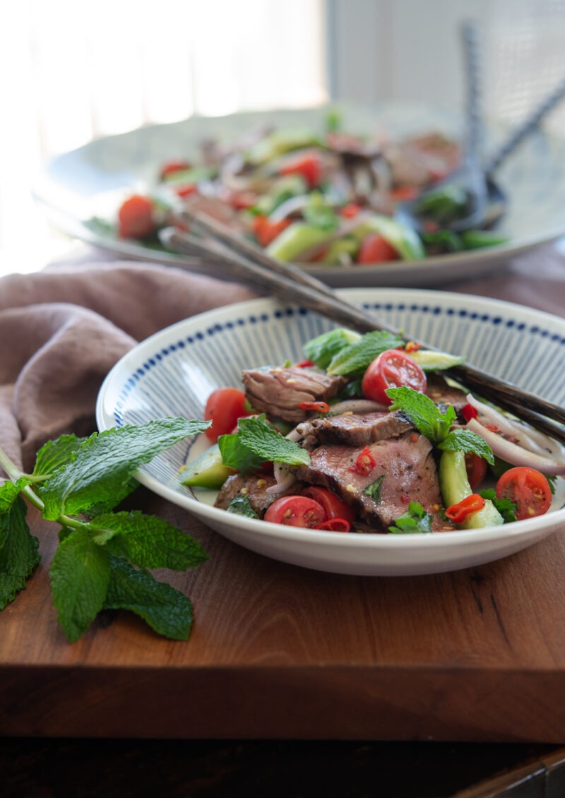 A plate of Thai beef salad is garnished with mint leaves and served with a pair of chopsticks