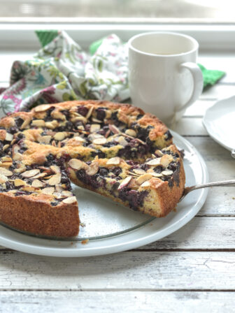 A slice of blueberry cake shows the burst of fresh blueberries on a white plate.