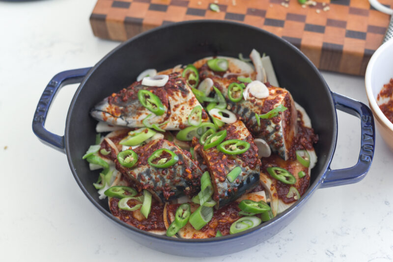 Sliced fresh green chilies are added on top of mackerel fish in a pot.
