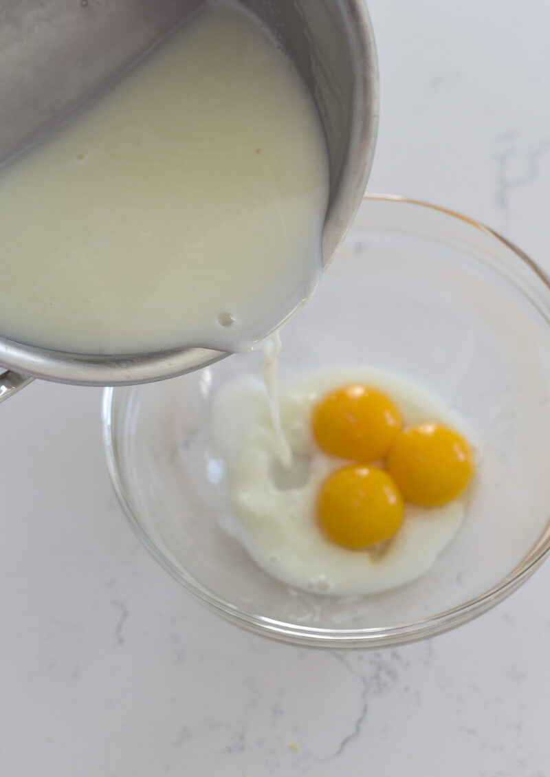 Hot lemon milk mixture is slowly poured on egg yolks in a bowl.