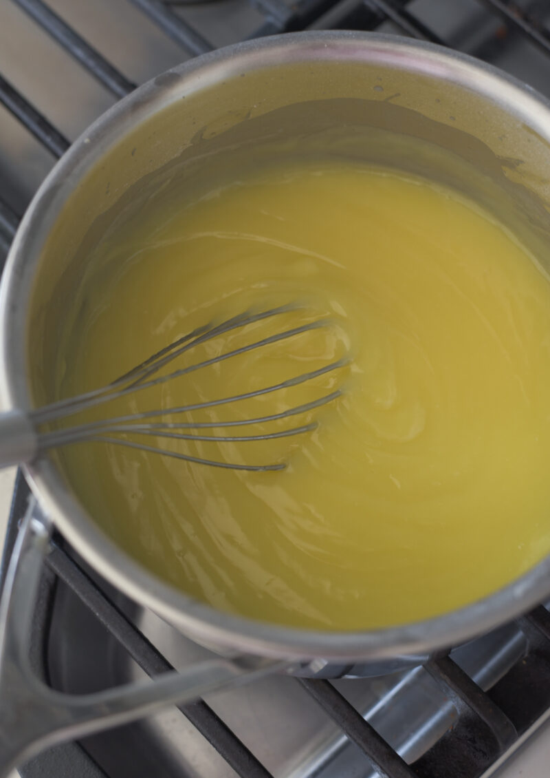 A whisk is stirring the lemon custard until thickened in a pan on the stove.