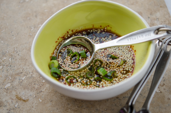 A soy dipping sauce with green onion and toasted sesame seeds are made in a small green bowl with a measuring spoon.