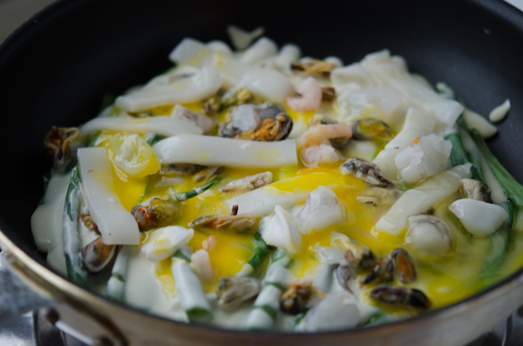 Beaten egg is drizzled over Korean seafood scallion pancake batter in a skillet.