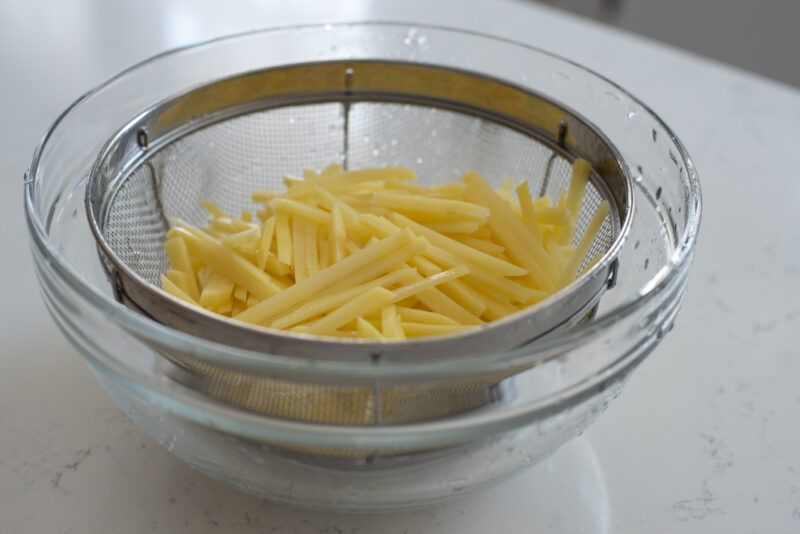 Potato strips are rinsed and drained in a colander inside a large bowl.
