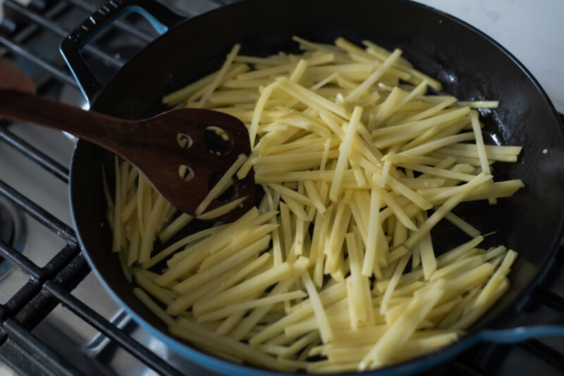 Potato strips are added to the skillet and a wooden spatula is stirring.
