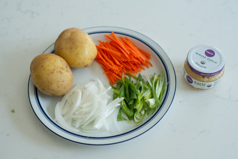 Two potatoes, slices of onion, carrot, and green onion are on a plate next to a jar of minced garlic.