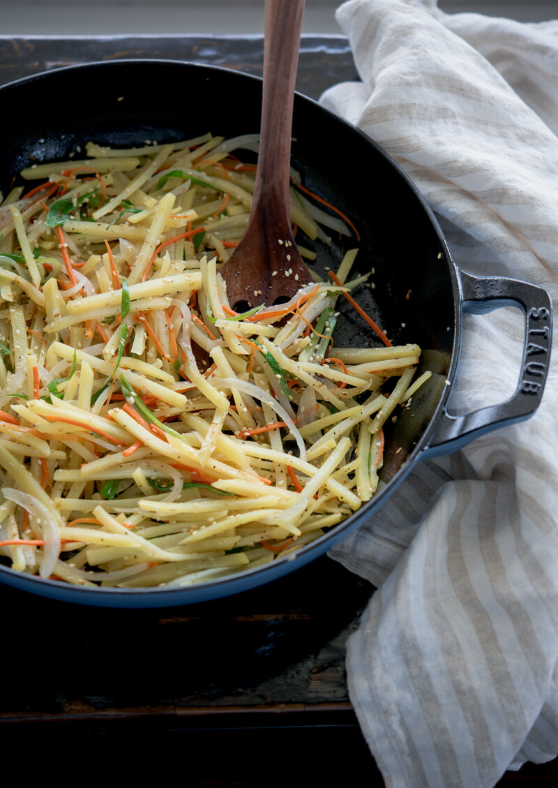Potato strips and shredded vegetables are pan fried in a pan with wooden spatula.