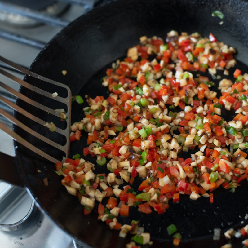 Mushroom, green onion, red pepper are cooking together with a spatula in a skillet
