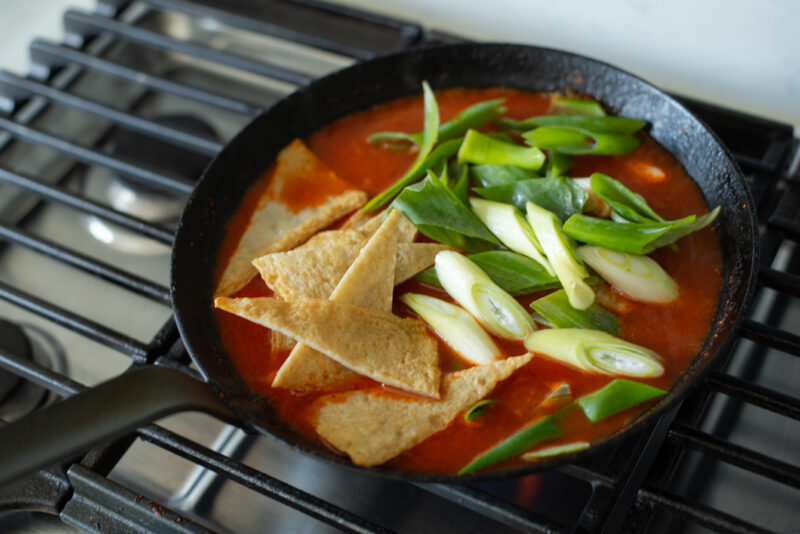 Fish cakes and leeks are added to simmering tteokbokki in a pan.