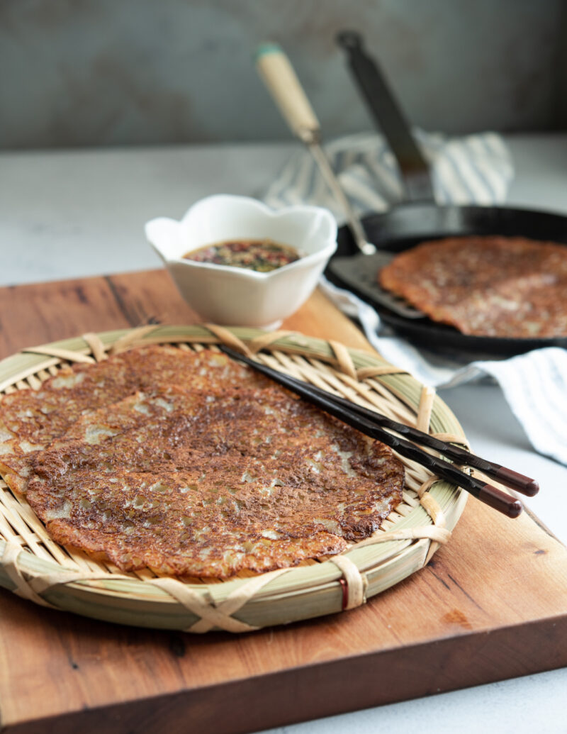 Golden crisp potato pancakes are served on a woven wooden plate with a pair of chopstick and soy chili dipping sauce.
