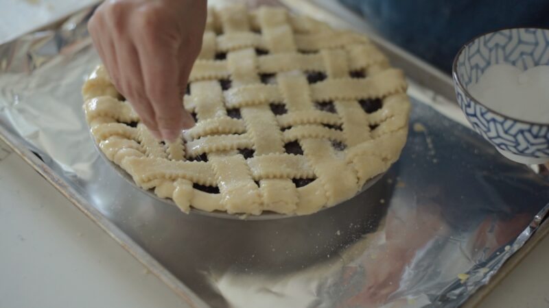 A hand is sprinkling sugar over the top pie crust.