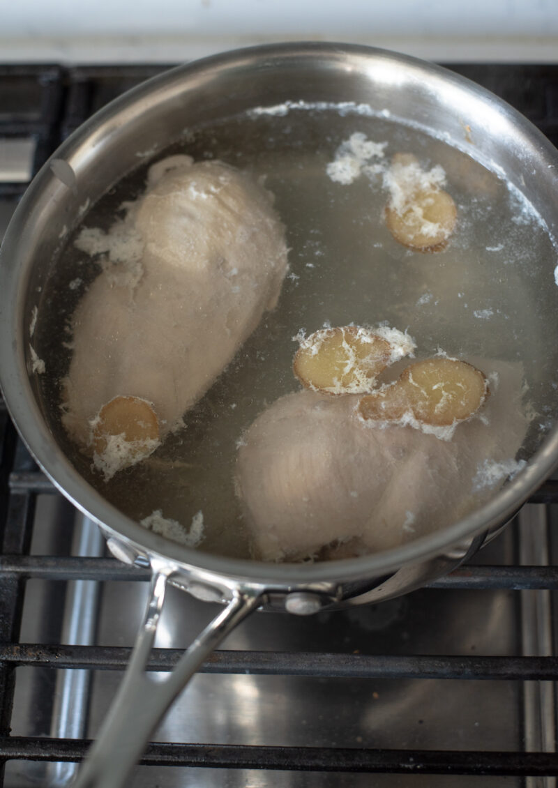 Two chicken breasts are poaching in a pot of simmering water with slices of ginger.
