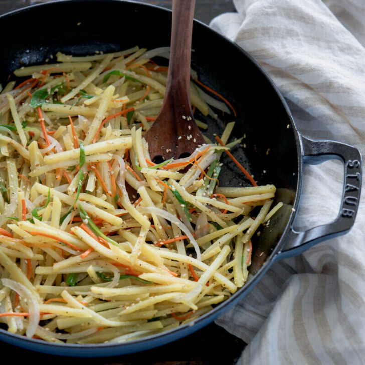 Potato strips and shredded vegetables are pan fried in a pan with wooden spatula.