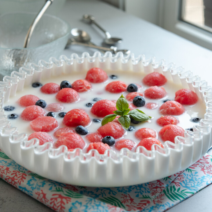 Watermelon balls are floating in a milky punch garnished with blueberries in a white bowl.