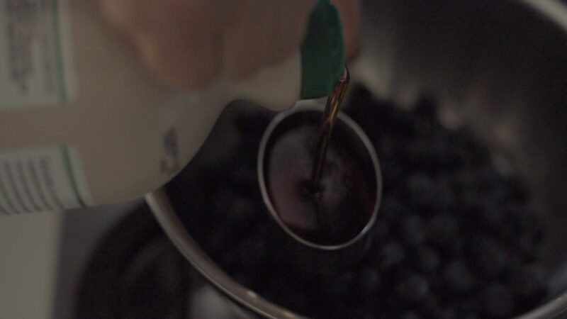 Maple syrup is measured in a cup and added to blueberries in a pot.
