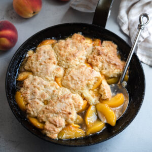 A skillet filled with biscuit topped peach cobbler is served with a large serving spoon.
