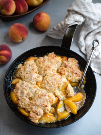 A skillet filled with biscuit topped peach cobbler is served with a large serving spoon.