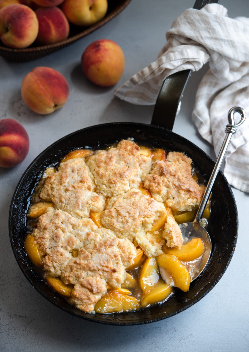 A skillet filled with biscuit topped peach cobbler is served with a large serving spoon.