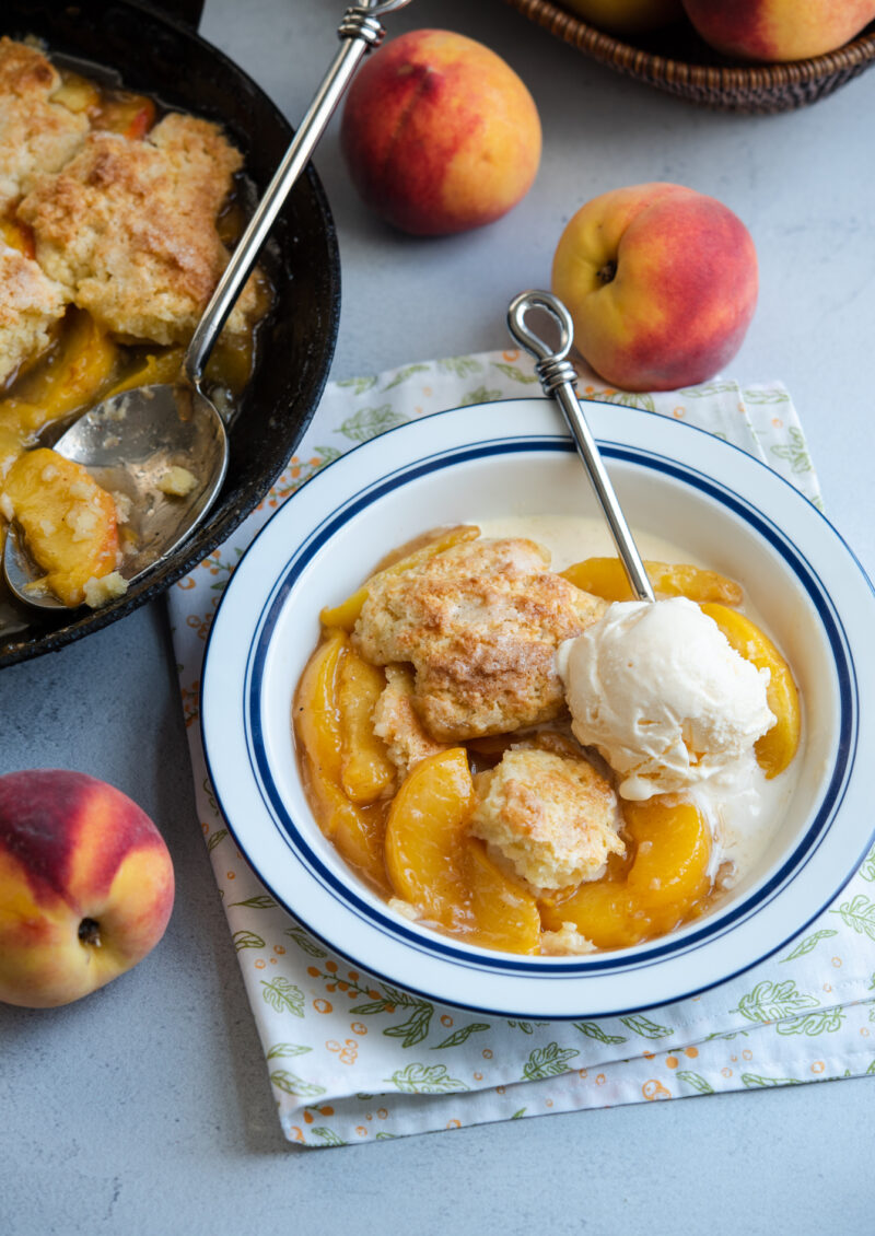 Old fashioned peach cobbler with biscuit topping is served with vanilla ice cream in a small serving bowl.