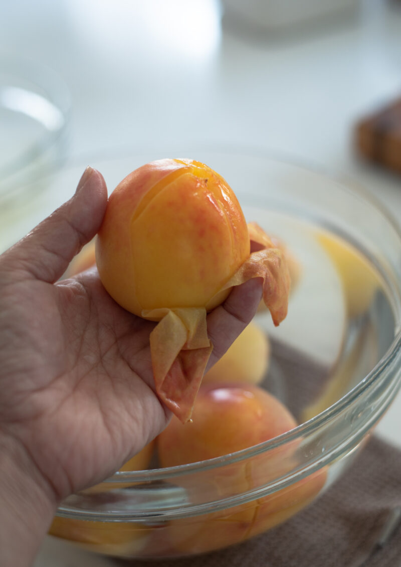 Skin of a peach that has been scored on the top is being peeled by hand revealing the juicy flesh underneath.
