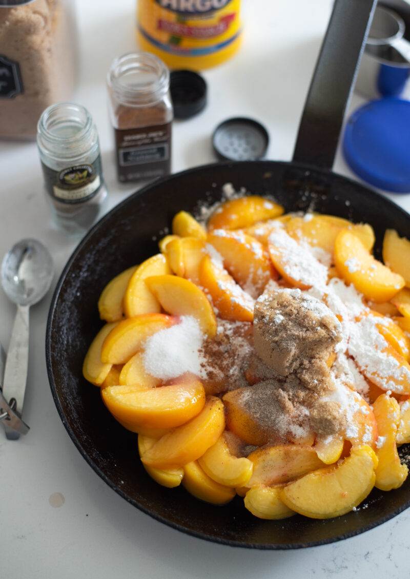 Brown sugar, white sugar, cornstarch are placed on top of fresh peach slices in a skillet.
