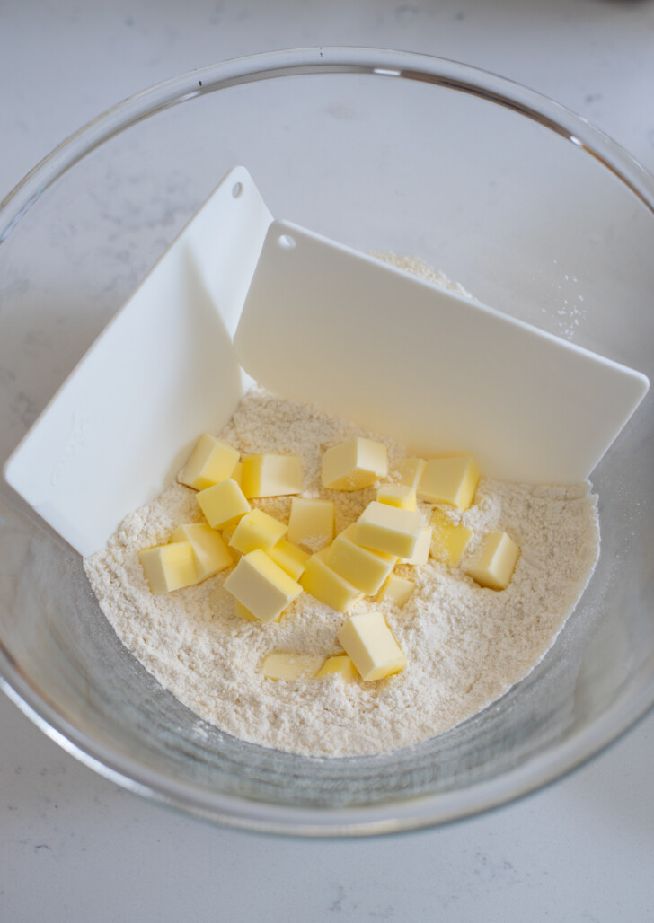 Small butter pieces are combined to flour mixture in a bowl with two dough scrapers.
