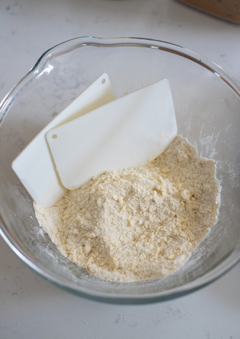 Two dough scrapers blended butter flour mixture into coarse crumbs in a bowl.