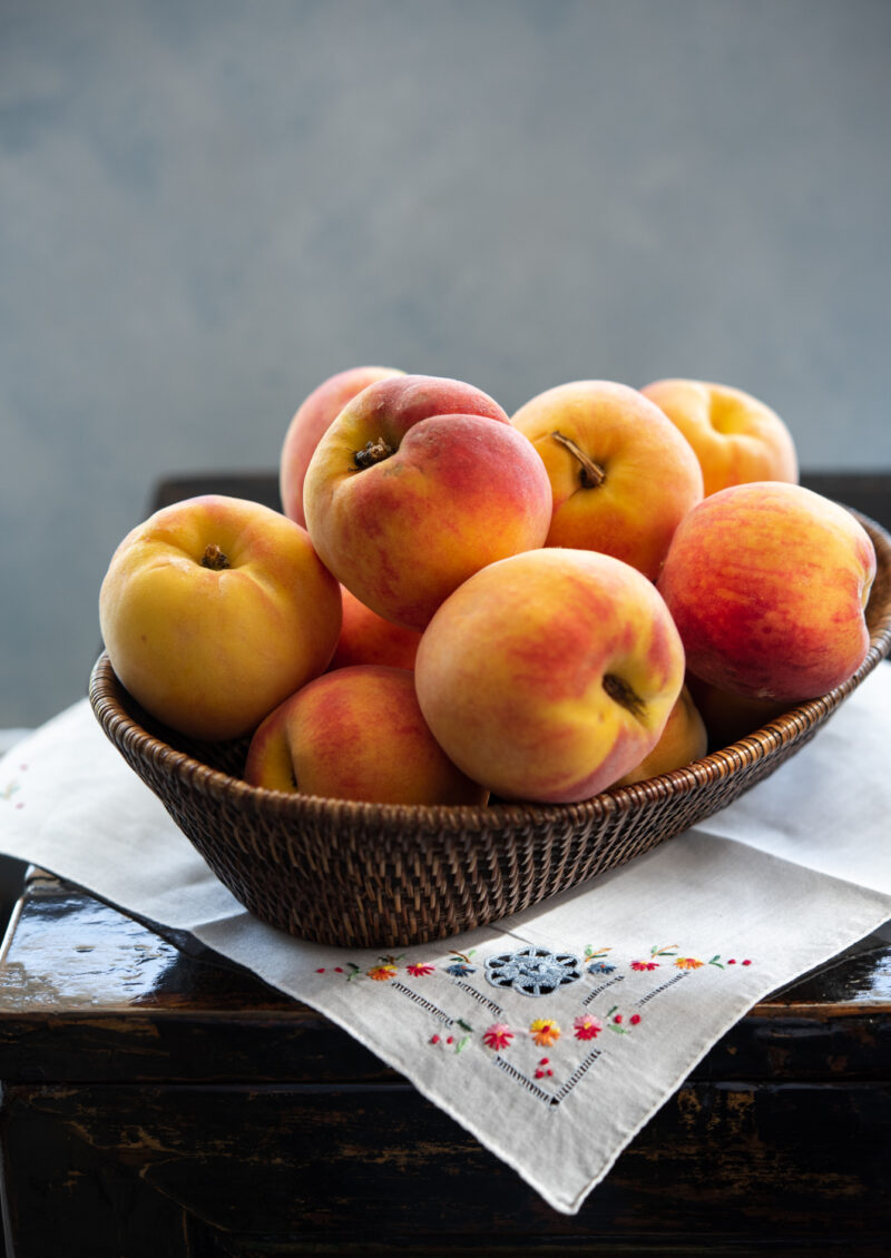 A woven basket filled with fresh peaches are placed on a embroidered napkin.