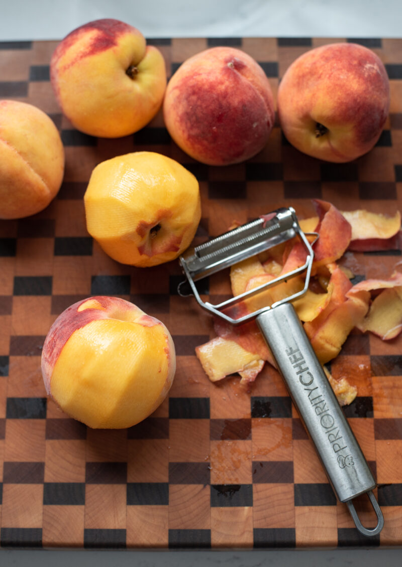 The skin of fresh peaches are being peeled by a vegetable peeler on the cutting board.