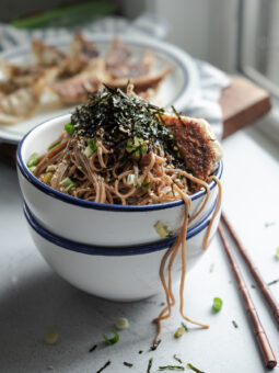 Sesame soba noodles are in a bowl topped with crumled seaweed and fried dumplings