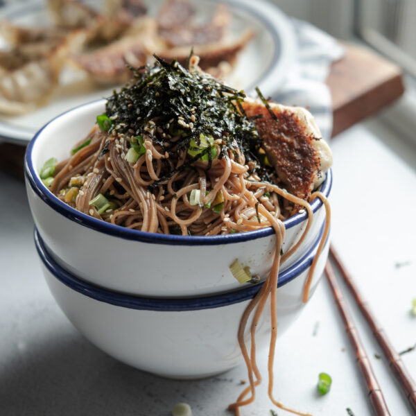 Sesame soba noodles are in a bowl topped with crumled seaweed and fried dumplings