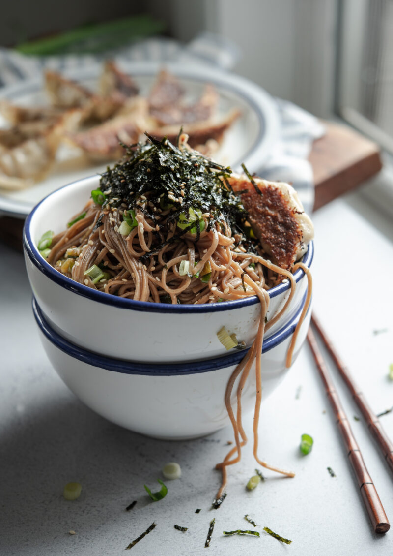 Sesame soba noodles are in a bowl topped with crumbled seaweed and fried dumplings