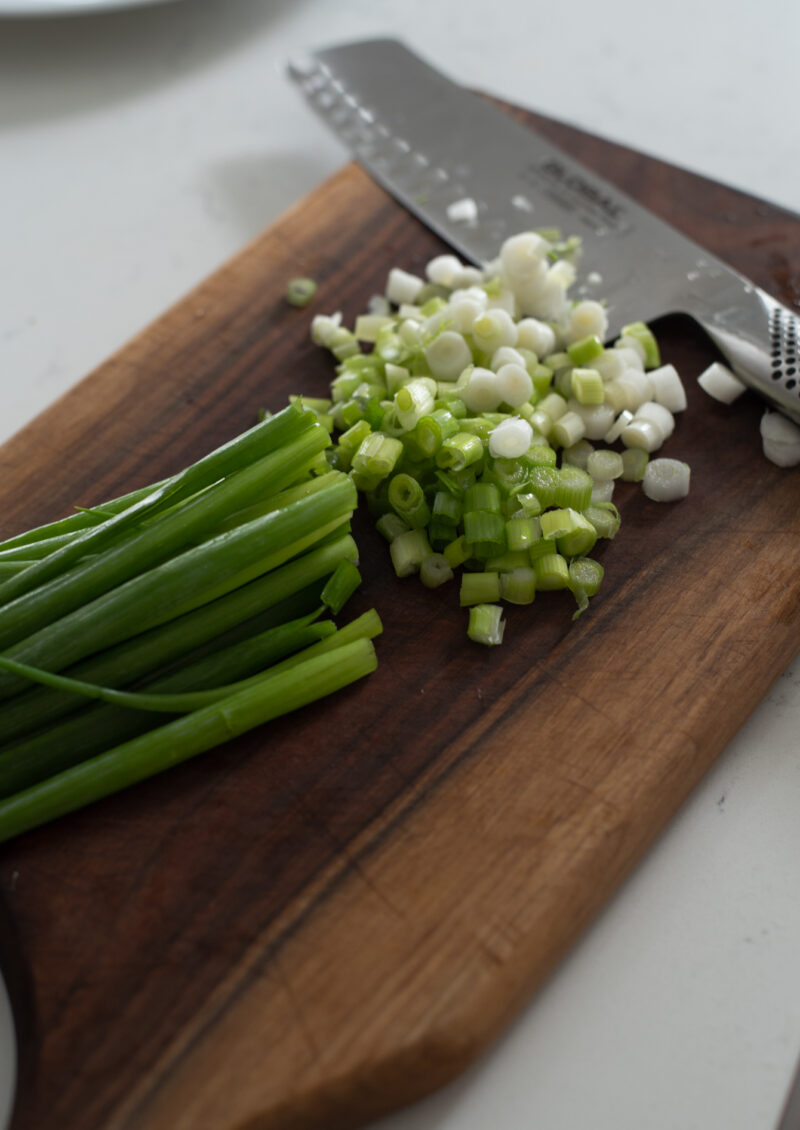 A bunch of green onion is chopped with a knife on the wooden cutting board.