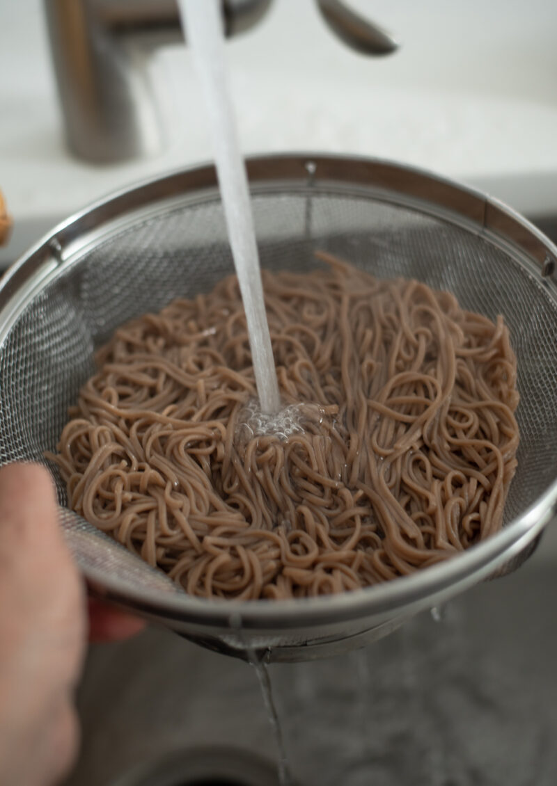 Cooked soba noodles in a colander are rinsing under the running water.