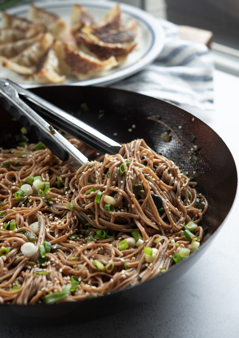 Kitchen tongs are holding sesame soba noodles garnished with green onion in a wok