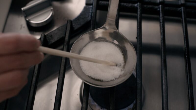 A wooden chopstick is stirring white sugar in a ladle over low heat on the stove.