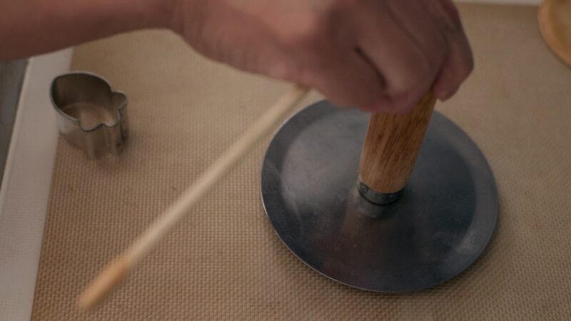 A flat metal round disk with a wooden handle is pressing down the dalgona candy.