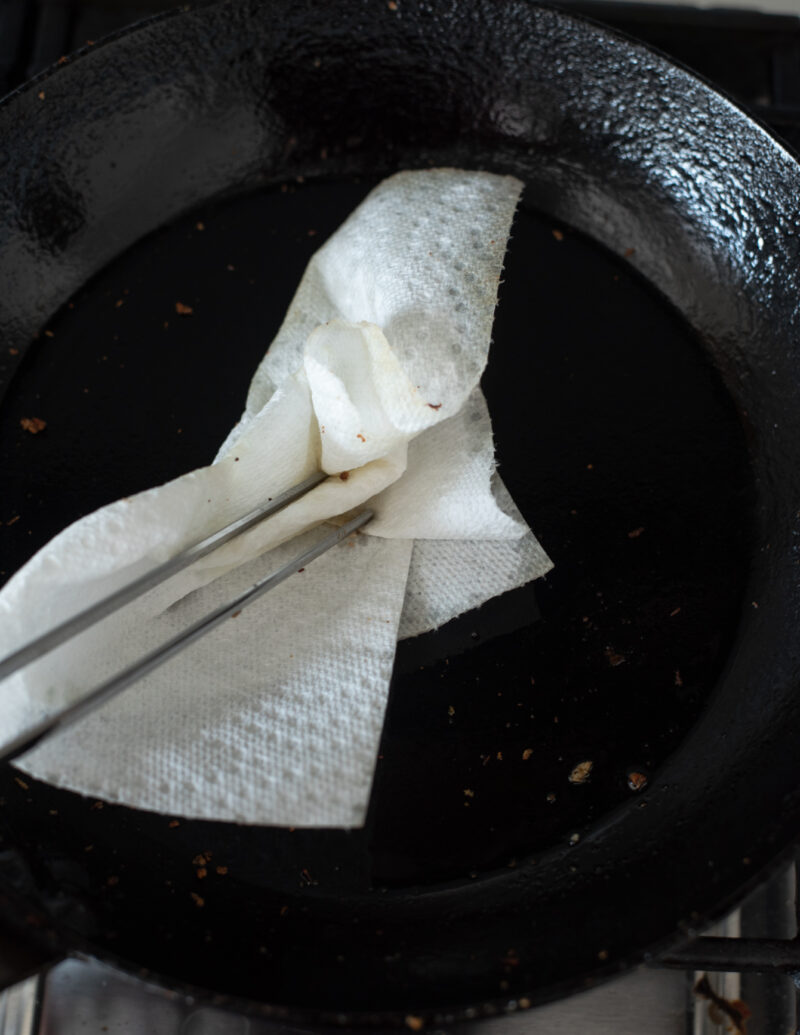 A kitchen tongs with paper towel is wiping out the excess oil from skillet.