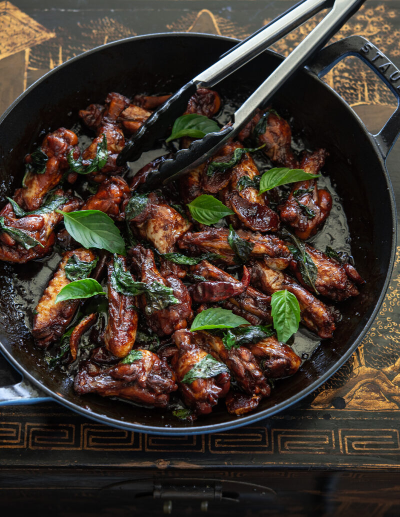 Three cup chicken wings (san bei ji) are adorned with fresh Thai basil leaves in a pan.