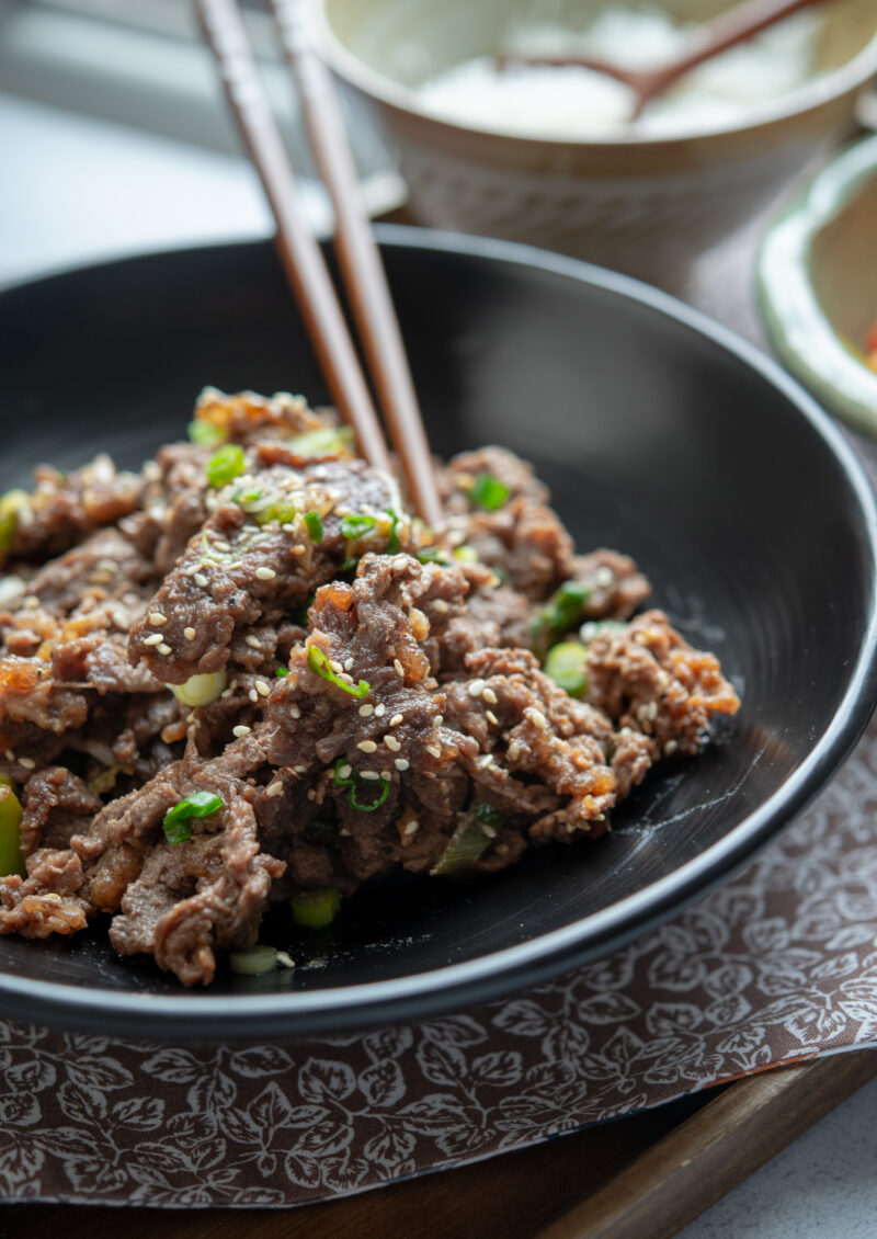 Tender Korean beef is garnished with green onion and served in a black bowl with chopsticks.